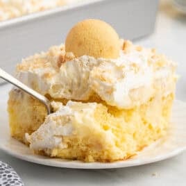 A slice of creamy yellow Banana Pudding Cake topped with whipped cream, crumbled cookies, and a whole vanilla wafer, served on a white plate with a fork.