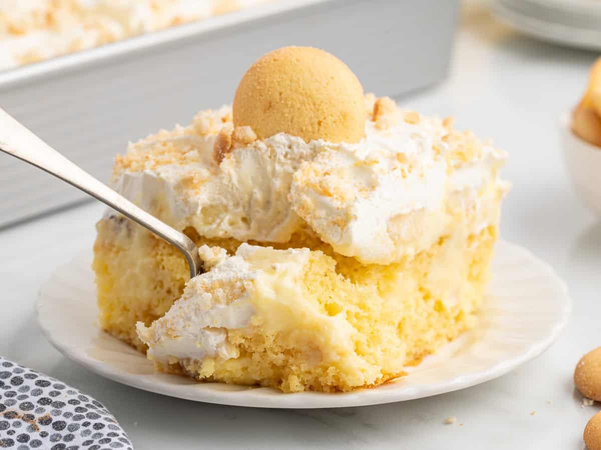 A slice of creamy yellow Banana Pudding Cake topped with whipped cream, crumbled cookies, and a whole vanilla wafer, served on a white plate with a fork.
