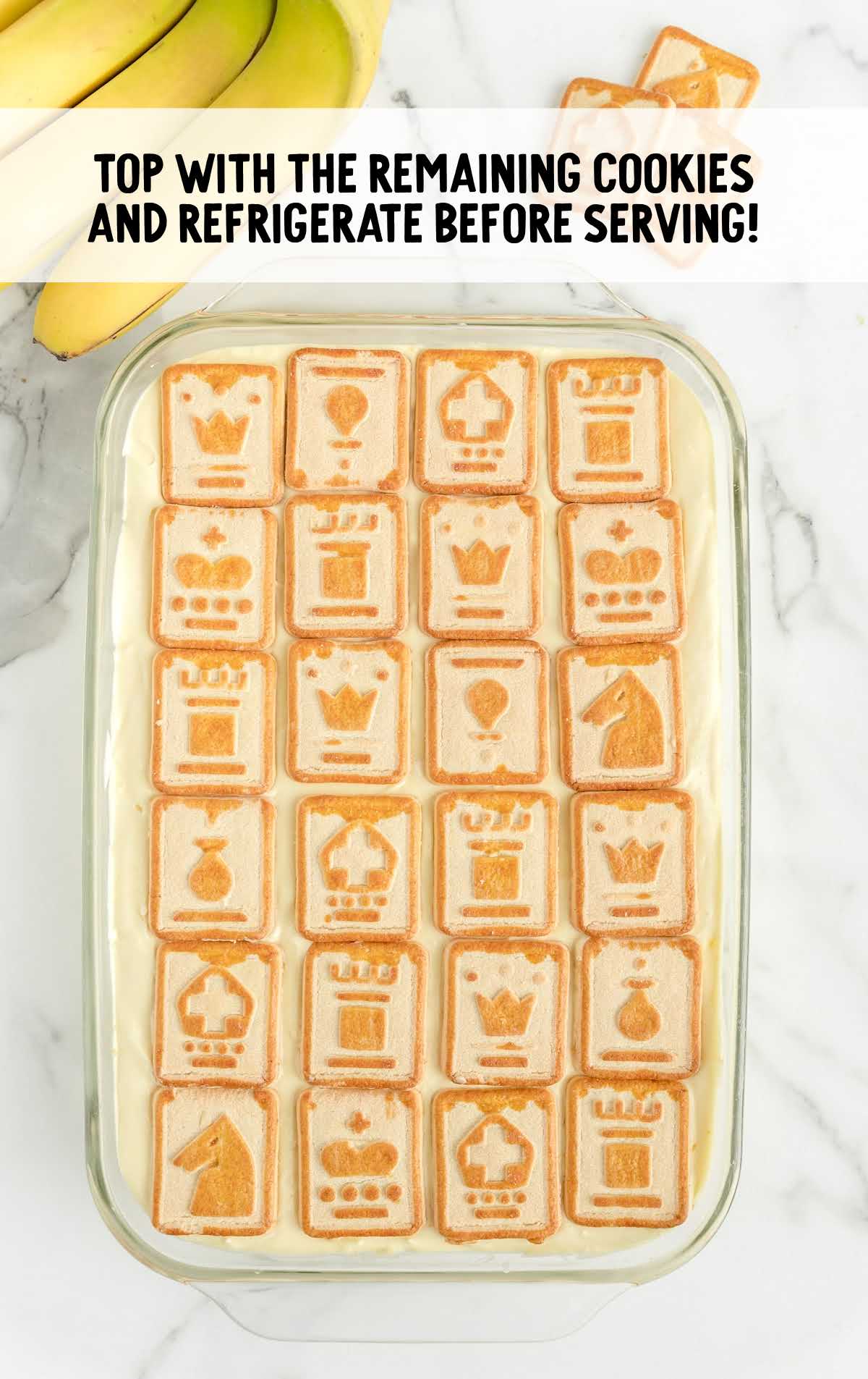 Overhead view of assembled chessmen banana pudding topped with a final layer of chessmen cookies in a glass dish, ready to refrigerate before serving.