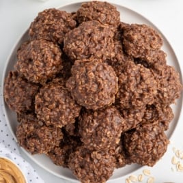 Overhead image of a white plate piled high with chocolate peanut butter no bake cookies made with oats, showing their rough, fudgy texture and rich chocolate color. A swirl of peanut butter and scattered oats are visible nearby, adding context to the ingredients.