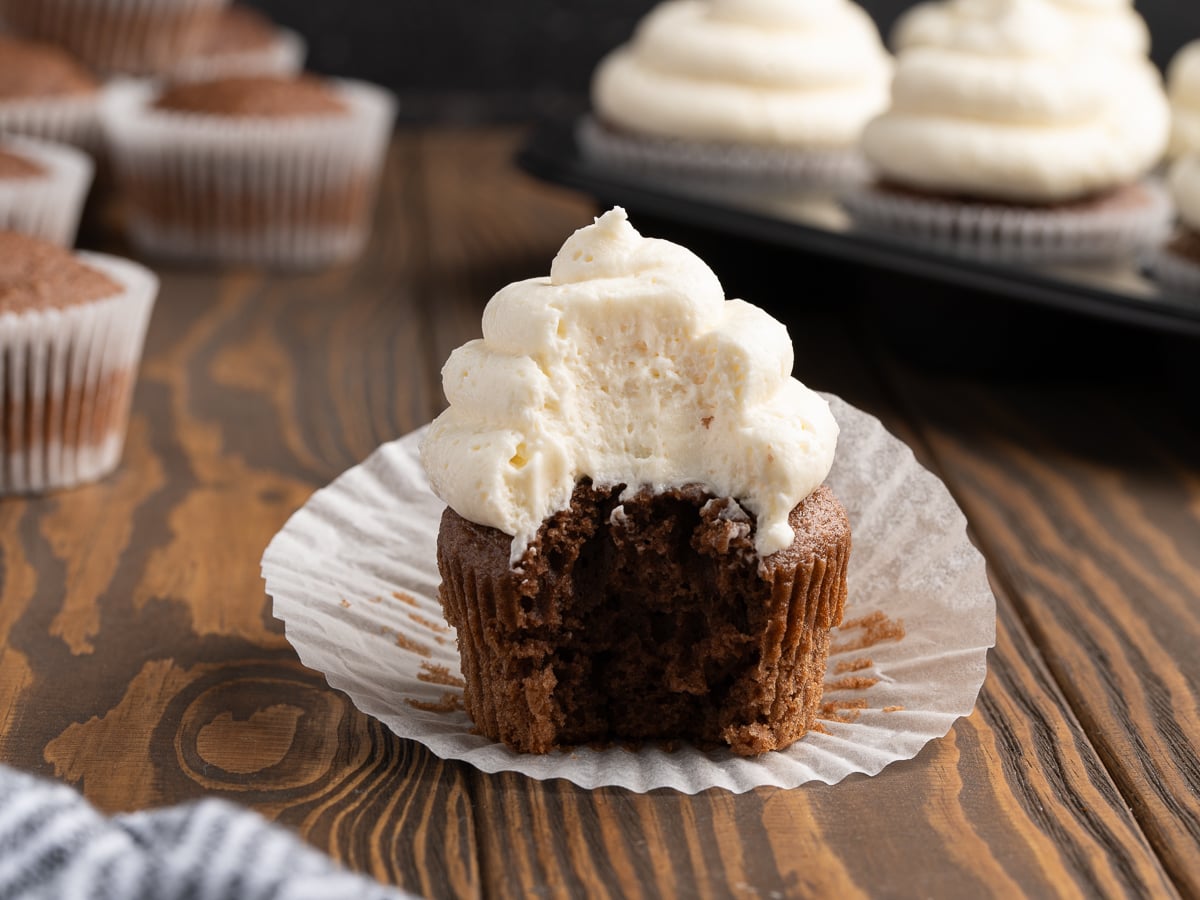 A chocolate cupcake with creamy white frosting, made from a Stabilized Whipped Cream Recipe, sits on an open wrapper with a bite taken out. More cupcakes with similar frosting are in the background on a wooden surface.