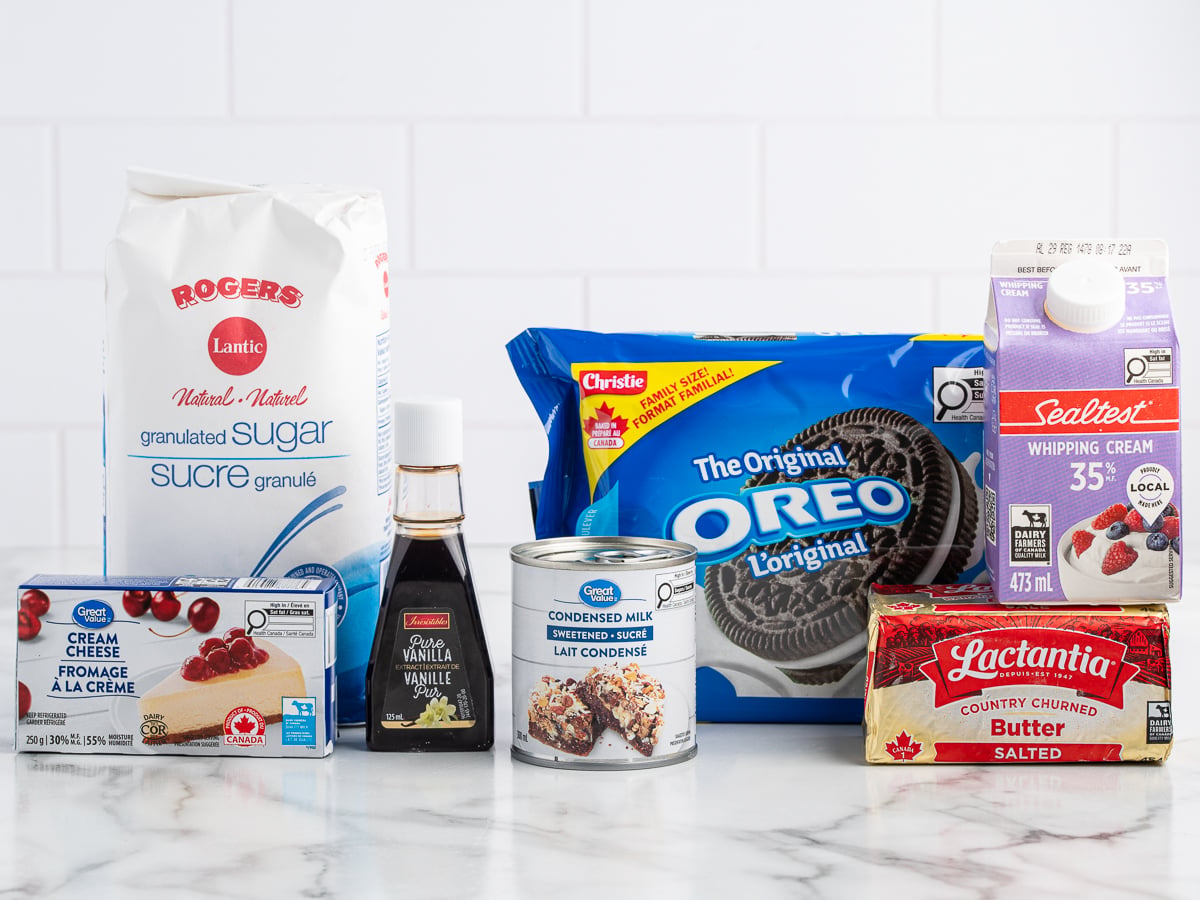 A variety of baking ingredients on a counter, perfect for making an Easy Frozen Oreo Dessert, including sugar, vanilla extract, cream cheese, condensed milk, Oreo cookies, whipping cream, and a stick of butter.