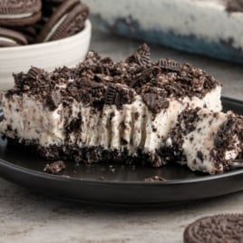 A close-up of a plate with a slice of Easy Frozen Oreo Dessert topped with crushed chocolate sandwich cookies. A fork has taken a bite, and a bowl of whole cookies is in the background.