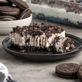 A large slice of Easy Frozen Oreo Dessert with a creamy Oreo filling and cookie crumble topping sits on a black plate, a bitten piece on a fork. A bowl of whole Oreos and a dessert dish complete this tempting treat in the background.
