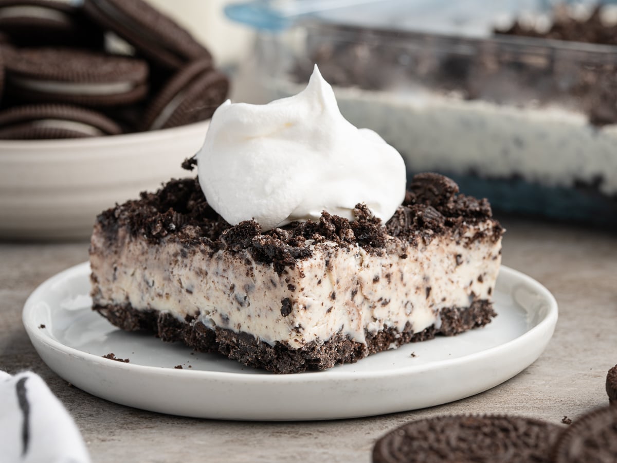 A square slice of creamy Easy Frozen Oreo Dessert topped with whipped cream, served on a white plate, with crushed cookies and a bowl of whole cookies in the background.