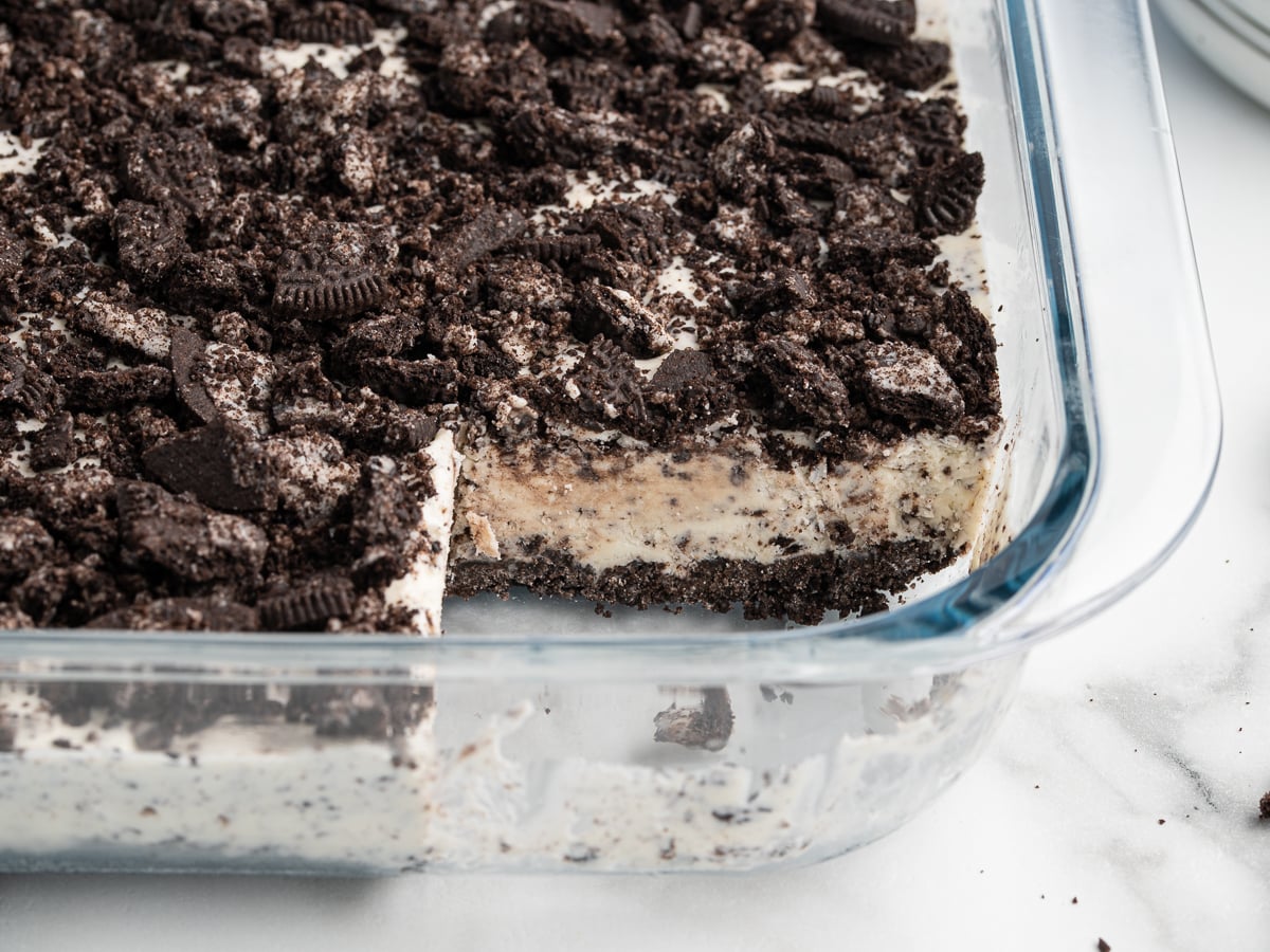 A glass baking dish filled with Easy Frozen Oreo Dessert, topped with crushed chocolate sandwich cookies, with a square piece removed to reveal the creamy layered treat inside.