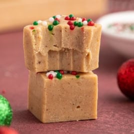Two square pieces of Gingerbread Fudge stacked, topped with red, green, and white sprinkles. The top piece has a bite taken out. Red and green glittery ornaments sit nearby on a reddish surface, adding festive flair to this irresistible Gingerbread Fudge Recipe.