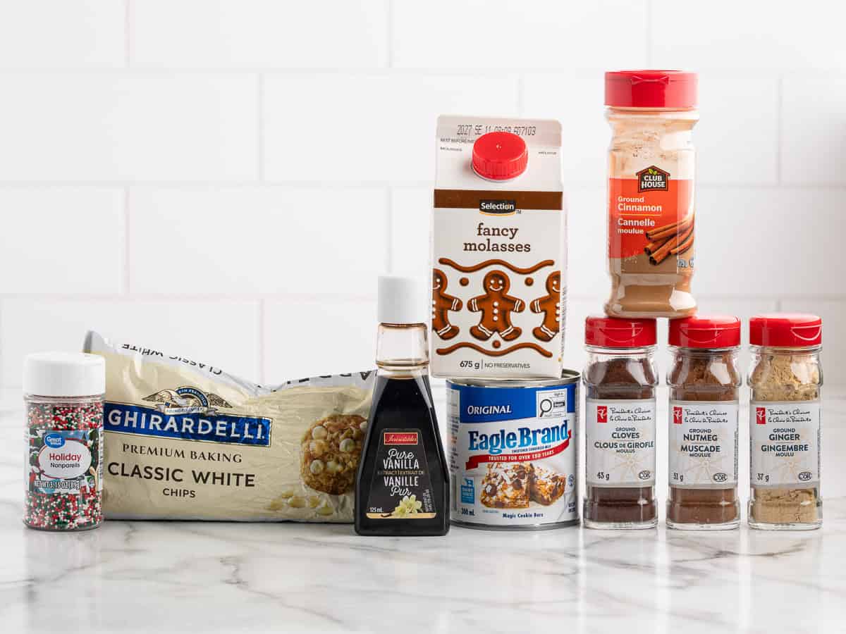 A selection of baking ingredients on a countertop, including white chocolate chips, vanilla extract, molasses, sprinkles, spices in jars, and a can of sweetened condensed milk—everything you need to create a delicious Gingerbread Fudge Recipe. White tile background.