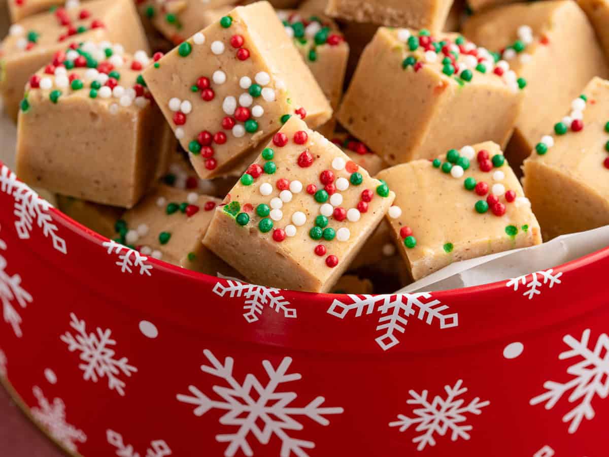 A red tin with white snowflakes holds decadent Gingerbread Fudge squares topped with red, green, and white sprinkles, suggesting a festive holiday treat.