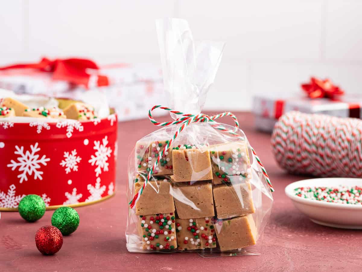 A clear bag of Gingerbread Fudge cubes topped with colorful sprinkles, tied with red and green twine, sits on a red table near a festive tin, Christmas ornaments, and a bowl of sprinkles—perfect for gifting or showcasing your favorite Gingerbread Fudge Recipe.
