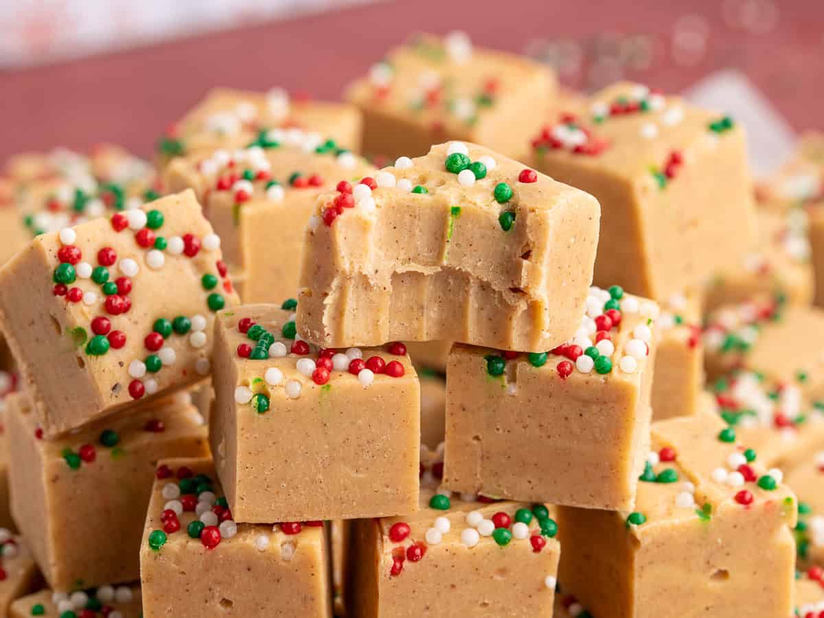 A pile of peanut butter fudge squares topped with red, green, and white sprinkles. The top piece has a bite taken out of it, revealing the creamy texture inside—perfect for serving alongside your favorite Gingerbread Fudge during the holidays.