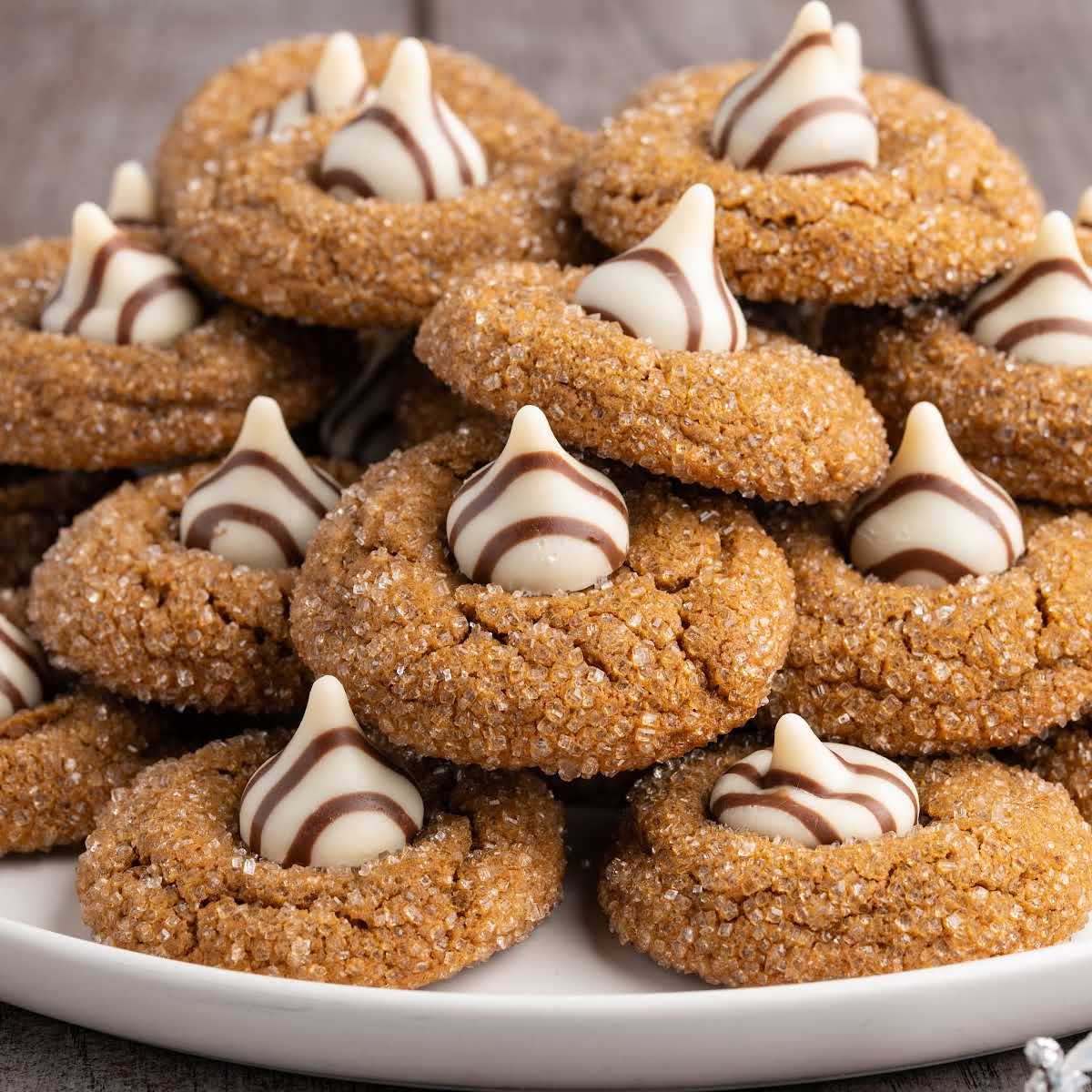 Pile of gingerbread kiss cookies coated in coarse sugar and topped with striped Hersheyโs Hugs chocolates on a white plate.
