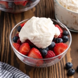 A glass bowl filled with fresh strawberries, blueberries, and raspberries, topped with a generous dollop of stabilized whipped cream, sits on a wooden table. Other bowls with cream and berries are in the background.