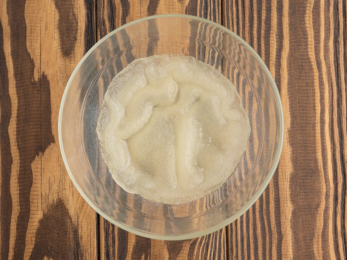 A glass bowl containing bloomed gelatin sits on a wooden surface, the gelatin appearing translucent and slightly grainy&mdash;perfect for making a stabilized whipped cream recipe or Homemade Cool Whip.