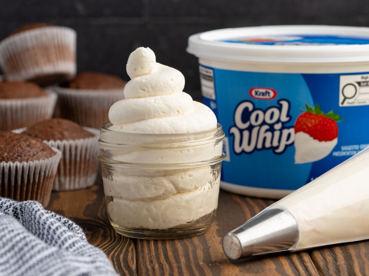 A glass jar filled with piped stabilized whipped cream sits on a wooden surface. A piping bag and a tub of Kraft Cool Whip are nearby, with chocolate cupcakes in the background.