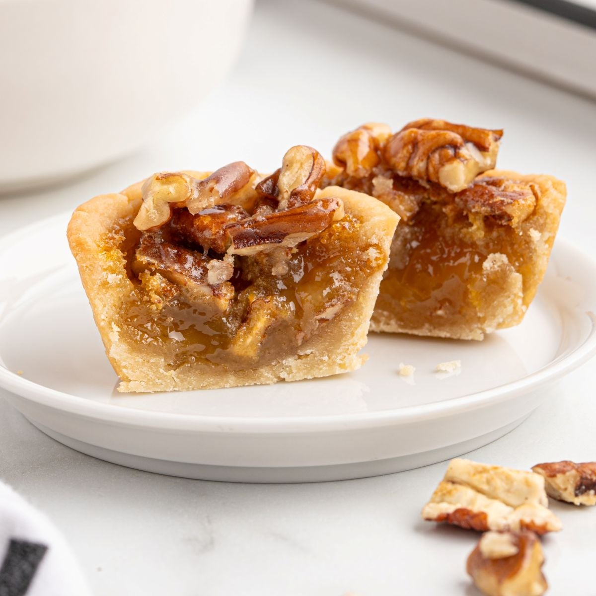 A close-up of a butter tart cut in half on a white plate, showing its gooey caramel filling and pecan topping, reminiscent of a Pecan Pie Bites Recipe. Crumbs and a few pecan pieces are scattered nearby on the plate.
