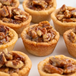 Mini Pecan Pie Bites with golden, flaky crusts, filled with gooey caramelized filling and topped with chopped pecans, arranged closely together on a white plate—an irresistible treat inspired by the classic Pecan Pie Bites Recipe.
