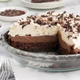 A delicious Mud Pie in a glass pie dish with a slice missing, showing layers of chocolate crust, creamy chocolate filling, and whipped topping with chocolate shavings. Plates and forks are in the background for this tempting Mud Pie recipe.