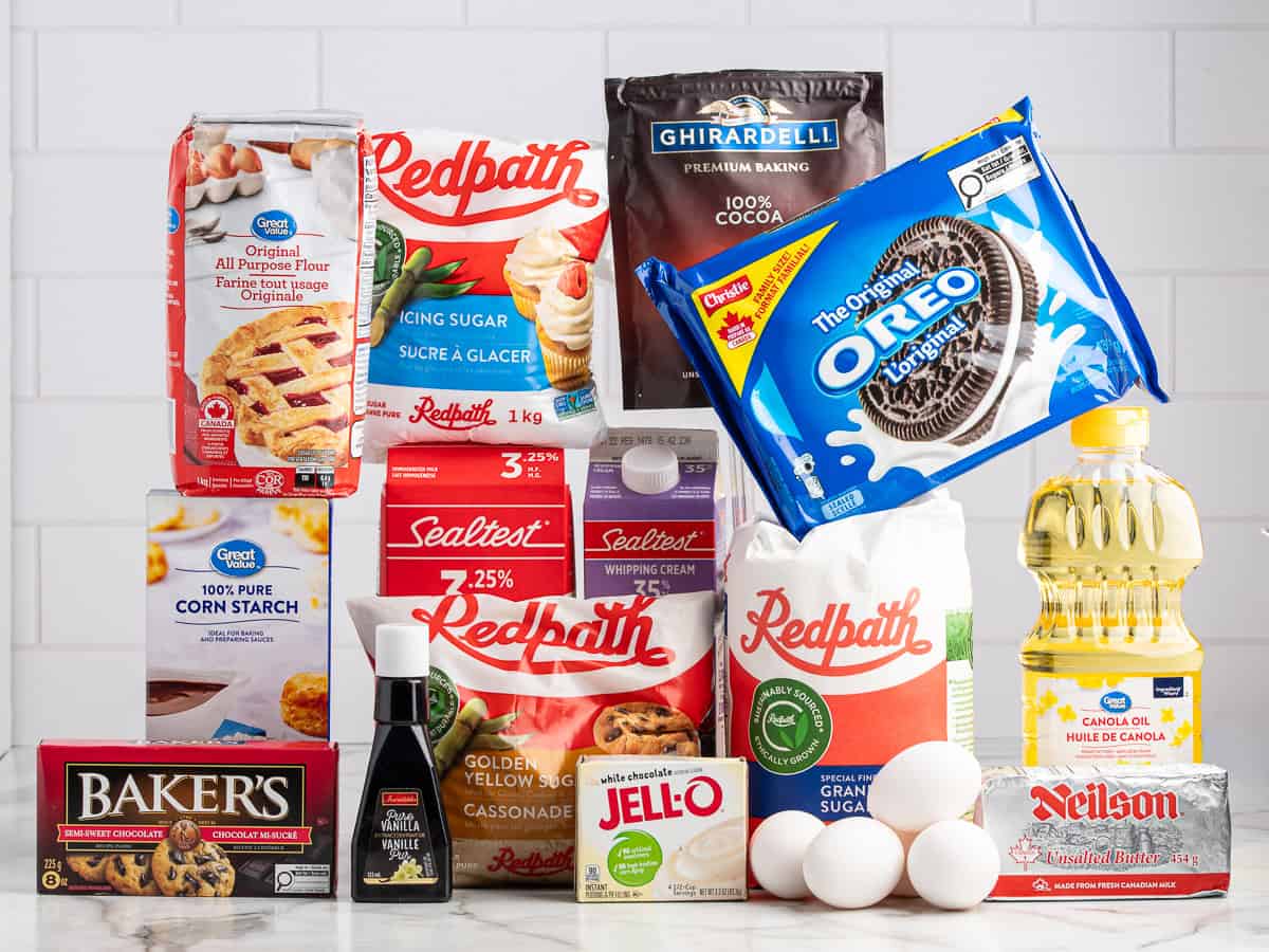 A variety of baking ingredients for a classic Mud Pie Recipe—including flour, sugar, cocoa powder, eggs, Oreos, Jello, and more—are arranged on a white countertop with a white tile background.