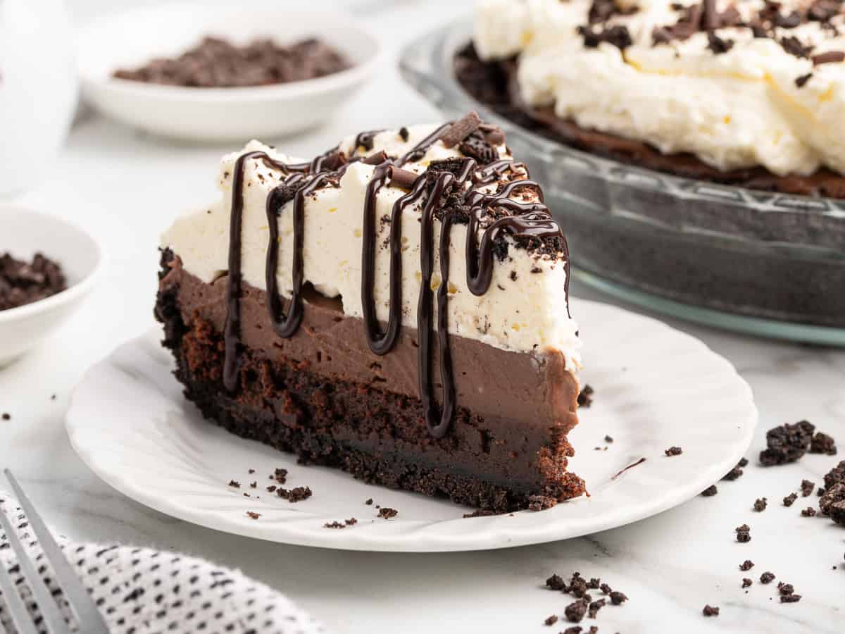 A slice of layered Mud Pie topped with whipped cream and chocolate drizzle sits on a white plate, with cookie crumbs scattered around and the rest of this irresistible Mud Pie Recipe visible in the background.