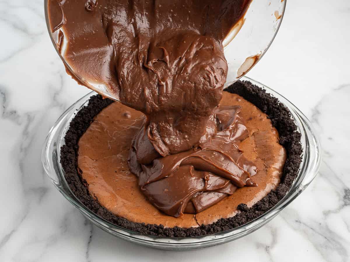 Thick chocolate filling is being poured from a glass bowl onto a partially filled Mud Pie with an Oreo cookie crust, on a white marble surface—the perfect moment in this Mud Pie Recipe.
