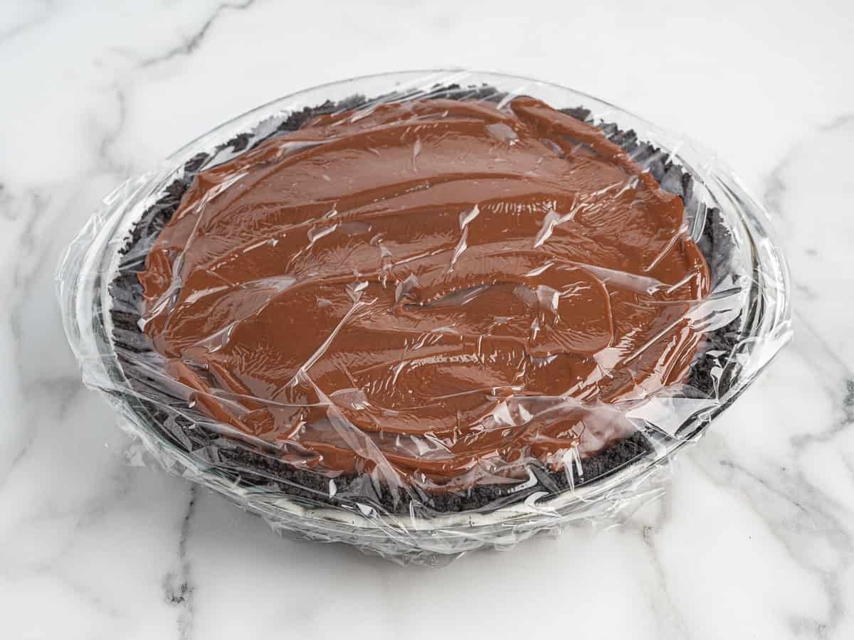 A Mud Pie with a dark cookie crust is covered in plastic wrap and sits on a white marble surface.
