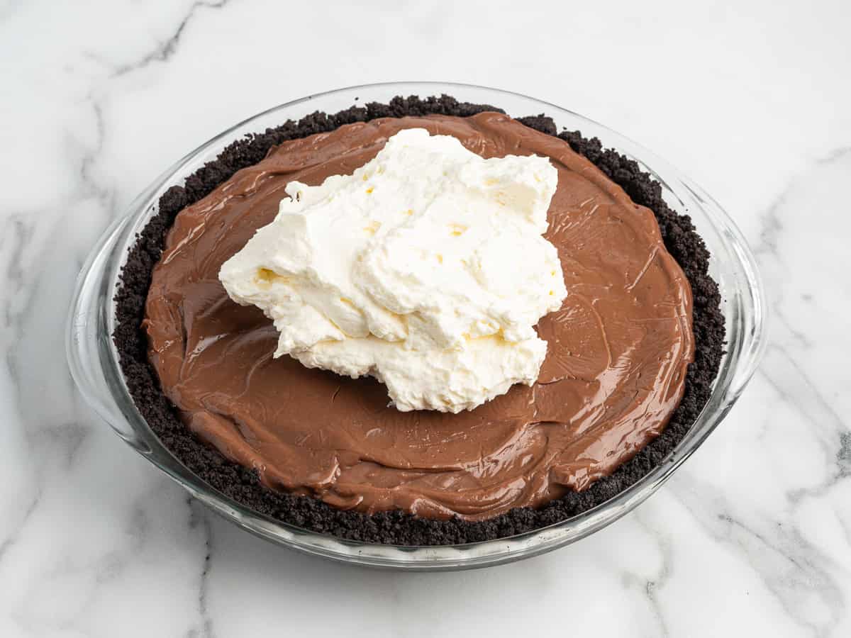 A decadent Mud Pie in a glass dish with an Oreo cookie crust, topped with a large dollop of whipped cream, rests on a white marble surface.