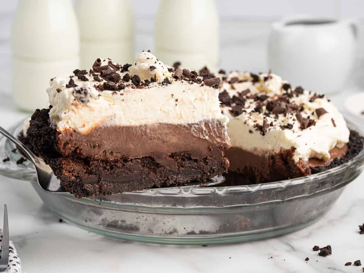 A rich Mud Pie with a dark cookie crust, chocolate layer, fluffy cream topping, and chocolate shavings sits on a marble counter as a slice is lifted from the pie plate. Milk bottles and a pitcher are in the background.