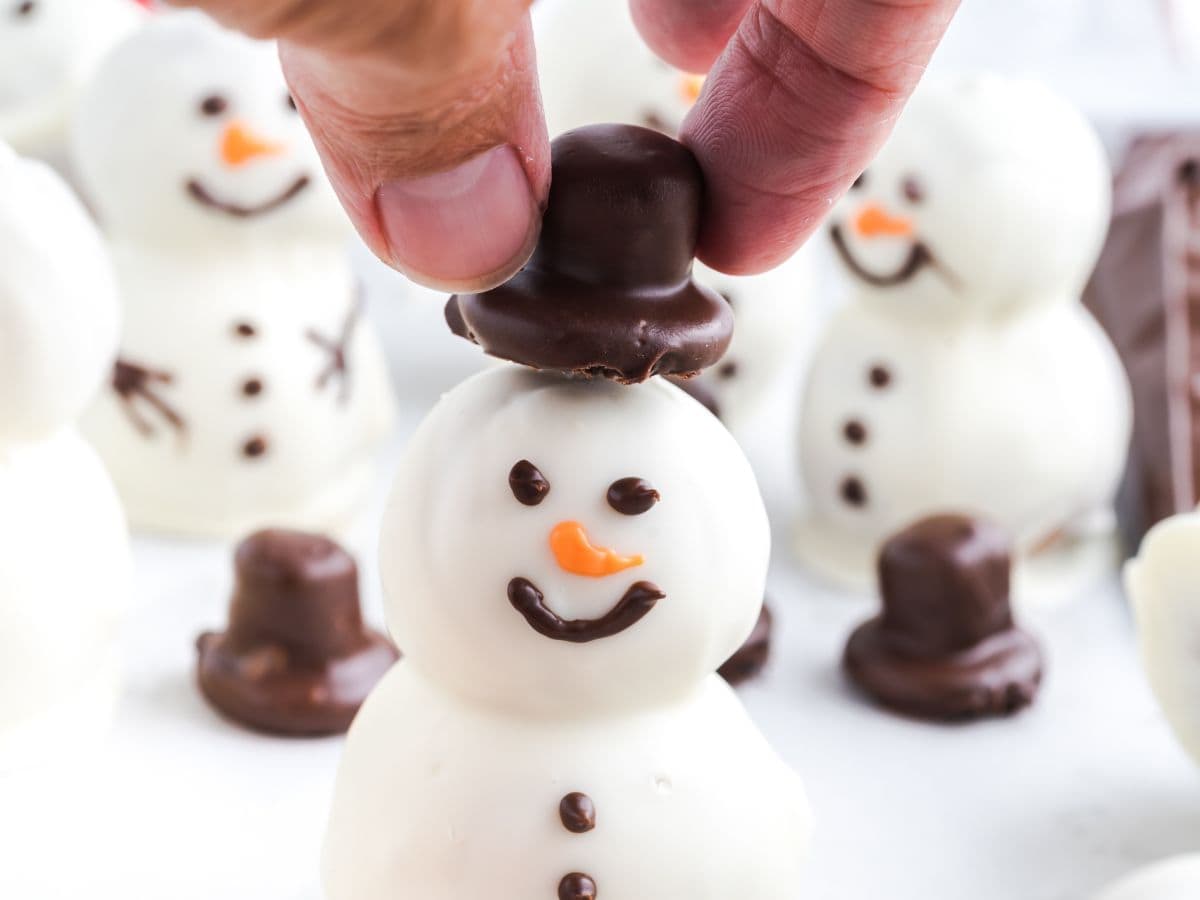 A hand places a chocolate hat on a snowman-shaped treat made of white chocolate with a smiling face, orange nose, and chocolate details—perfect for any Snowmen Truffles Recipe. Other similar Snowmen Truffles are visible in the background.