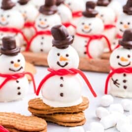 Cute snowmen truffles made from white chocolate, with chocolate hats, candy scarves, and cookie bases, are arranged on a table with mini marshmallows and biscuits. Some snowmen are grouped on a wooden tray in the background, making this Snowmen Truffles Recipe perfect for holiday entertaining.