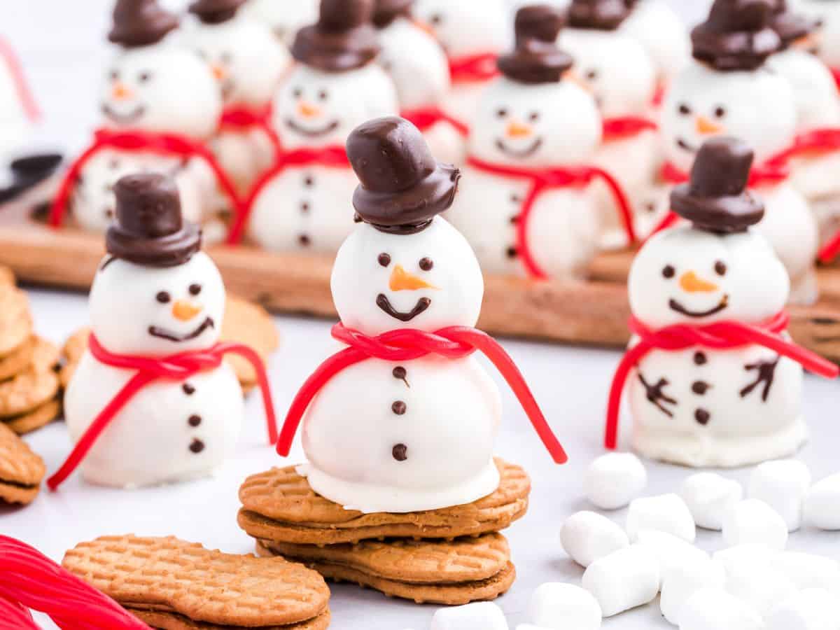 Cute snowmen truffles made from white chocolate, with chocolate hats, candy scarves, and cookie bases, are arranged on a table with mini marshmallows and biscuits. Some snowmen are grouped on a wooden tray in the background, making this Snowmen Truffles Recipe perfect for holiday entertaining.