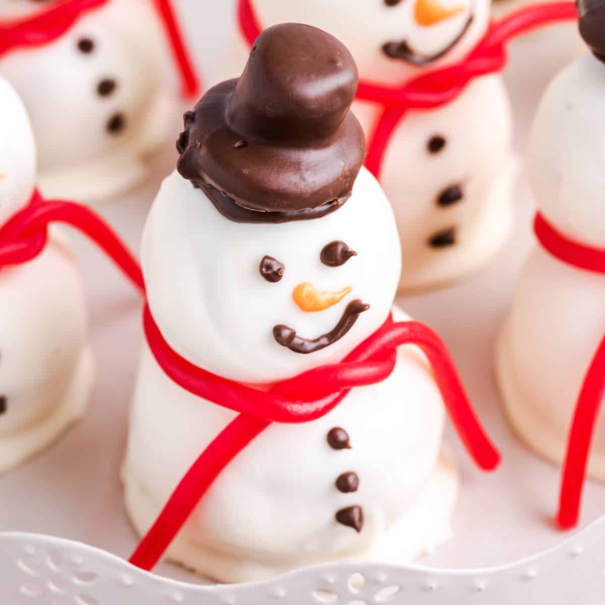 A close-up of a white chocolate-covered snowman treat from the Snowmen Truffles Recipe, featuring chocolate eyes, buttons, and hat, an orange nose, and a red candy scarf. Other similar Snowmen Truffles are visible in the background.