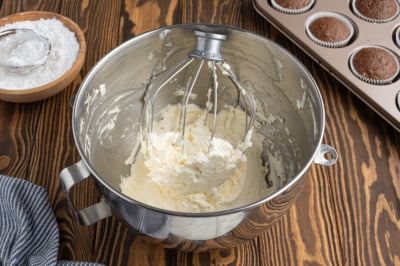 A metal mixing bowl with a whisk attachment holds homemade Cool Whip-style frosting. Nearby are a bowl of powdered sugar, a striped towel, and a muffin tin with chocolate cupcakes on a wooden surface.