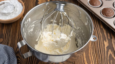 A metal mixing bowl with a whisk attachment holds homemade Cool Whip-style frosting. Nearby are a bowl of powdered sugar, a striped towel, and a muffin tin with chocolate cupcakes on a wooden surface.