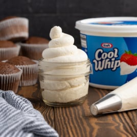 A glass jar filled with swirled stabilized whipped cream sits on a wooden surface next to a piping bag, a container of Cool Whip, several chocolate cupcakes in paper liners, and a striped kitchen towel.