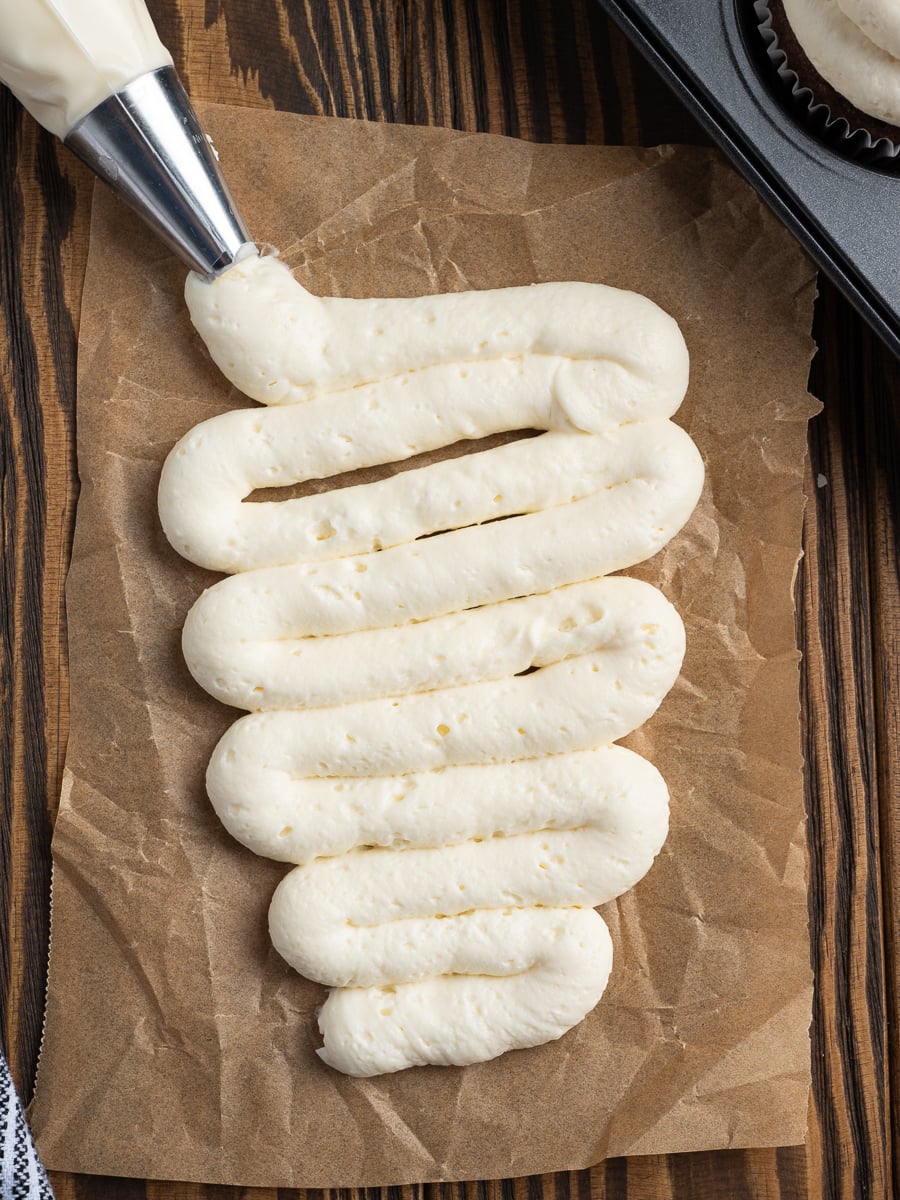 Thick, creamy white frosting&mdash;resembling a Homemade Cool Whip&mdash;is being piped in a zigzag pattern onto a sheet of brown parchment paper, with a metal piping tip visible at the top left.