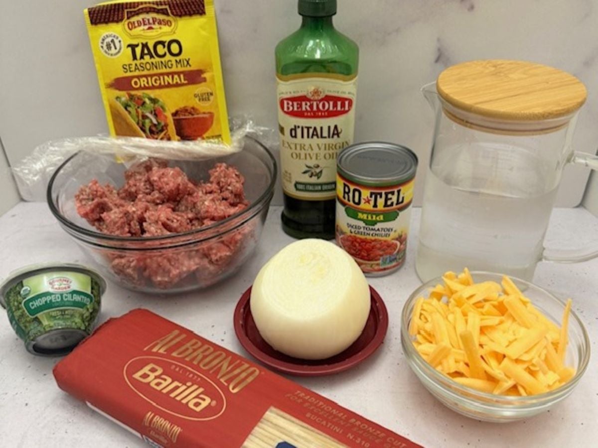 A variety of ingredients on a counter for a Taco Spaghetti Recipe: ground beef, taco seasoning, olive oil, tomatoes with green chilies, chopped cilantro, white onion, shredded cheddar cheese, spaghetti, and a pitcher of water.