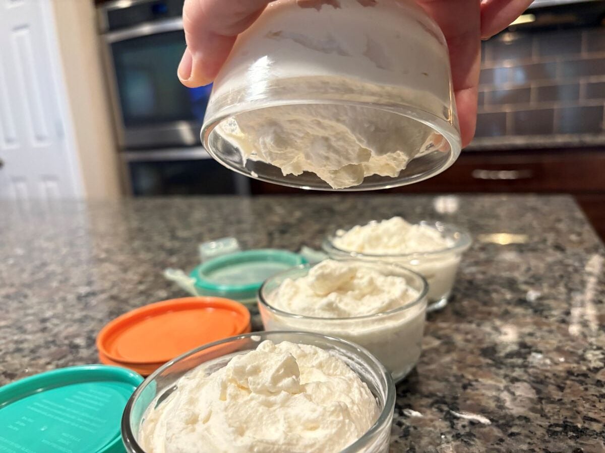 A hand holds a small glass container of whipped cream cheese above a countertop, with three similar containers&mdash;perfect for storing your Homemade Cool Whip or Stabilized Whipped Cream Recipe&mdash;in a cozy kitchen setting.