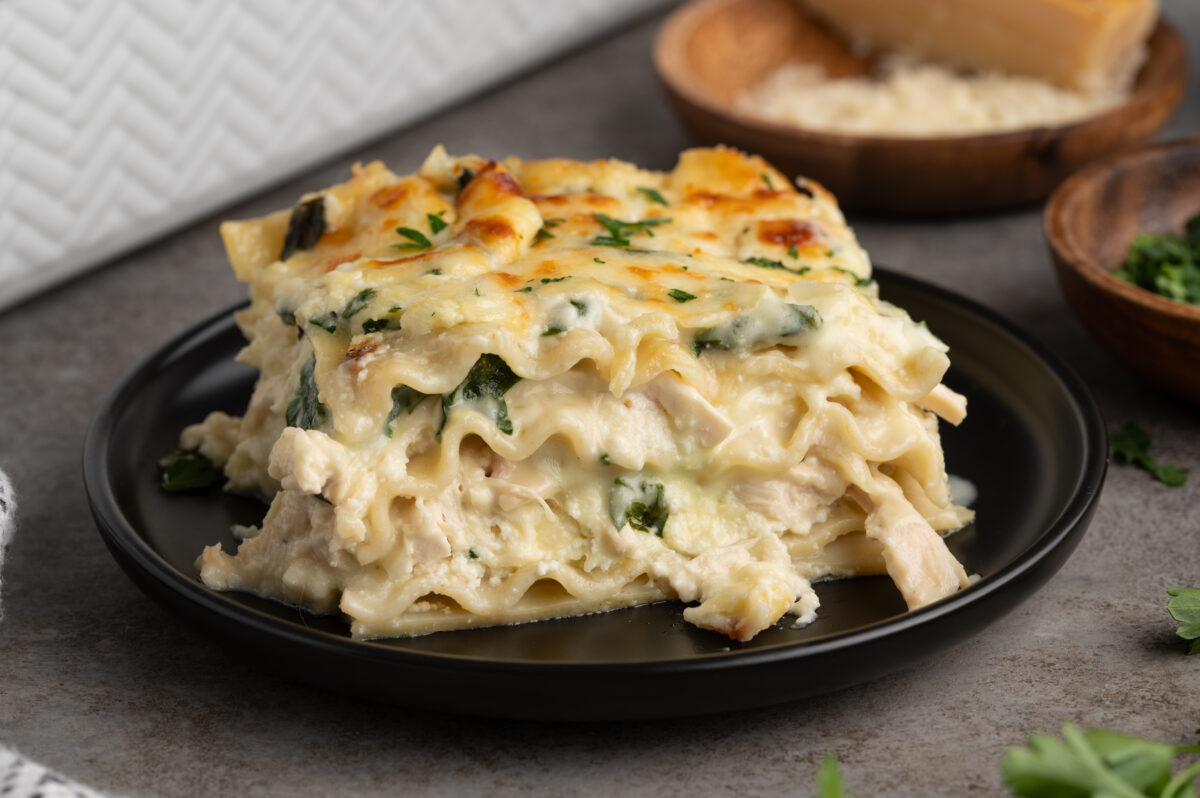 A slice of creamy chicken lasagna with spinach, topped with melted cheese and parsley, is served on a black plate, with bowls of grated cheese and herbs in the background&mdash;perfect for your favorite chicken lasagna recipe.