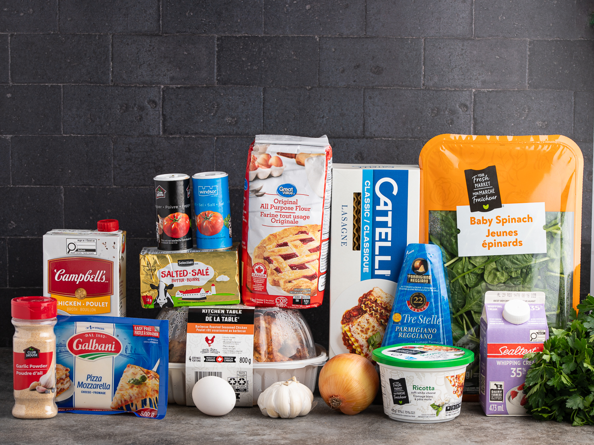 A variety of lasagna ingredients for a delicious white chicken lasagna recipe are arranged on a kitchen counter, including pasta, spinach, cheeses, tomato products, chicken broth, eggs, onion, garlic, spices, and herbs against a dark brick background.