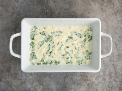 A white rectangular baking dish with handles contains a layer of creamy mixture and visible green spinach pieces, perfect for a white chicken lasagna recipe, set on a gray textured surface.