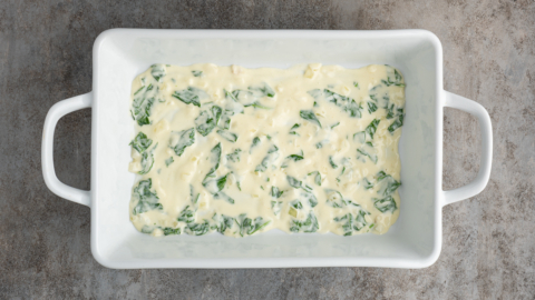 A white rectangular baking dish with handles contains a layer of creamy mixture and visible green spinach pieces, perfect for a white chicken lasagna recipe, set on a gray textured surface.