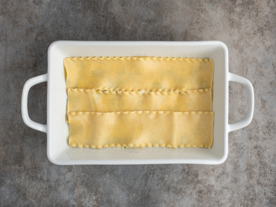 A white rectangular baking dish with three uncooked lasagna noodles laid flat side by side on the bottom, ready to become a delicious white chicken lasagna recipe, set on a textured gray surface.