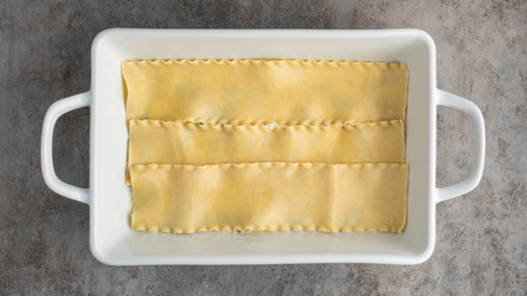 A white rectangular baking dish with three uncooked lasagna noodles laid flat side by side on the bottom, ready to become a delicious white chicken lasagna recipe, set on a textured gray surface.