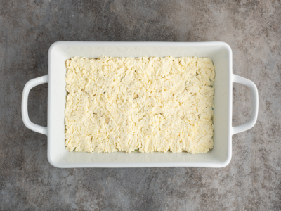 A white rectangular baking dish filled with an even layer of yellowish, crumbly mixture—perfect for a white lasagna recipe—sits on a mottled gray countertop.