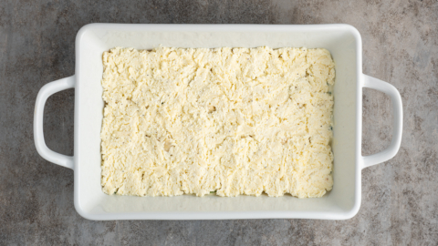 A white rectangular baking dish filled with an even layer of yellowish, crumbly mixture&mdash;perfect for a white lasagna recipe&mdash;sits on a mottled gray countertop.