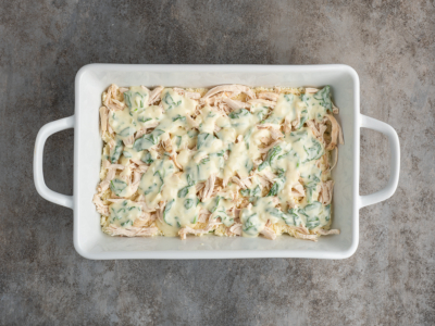 A white rectangular baking dish filled with shredded chicken topped with a creamy spinach sauce, resembling a delicious white chicken lasagna recipe, placed on a textured gray surface.