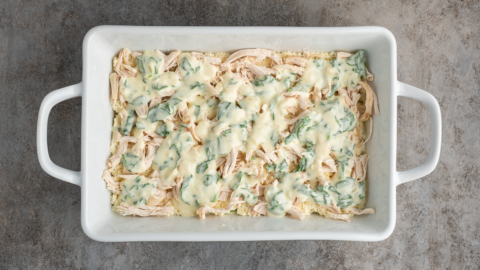 A white rectangular baking dish filled with shredded chicken topped with a creamy spinach sauce, resembling a delicious white chicken lasagna recipe, placed on a textured gray surface.