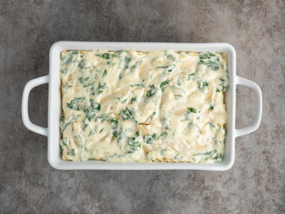 A rectangular white baking dish filled with an uncooked lasagna with cream cheese and spinach, topped with a creamy white sauce, sits on a gray textured surface.