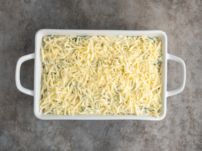 A rectangular white baking dish filled with an unbaked white chicken lasagna recipe, topped with a generous layer of shredded cheese, sits on a gray textured surface.